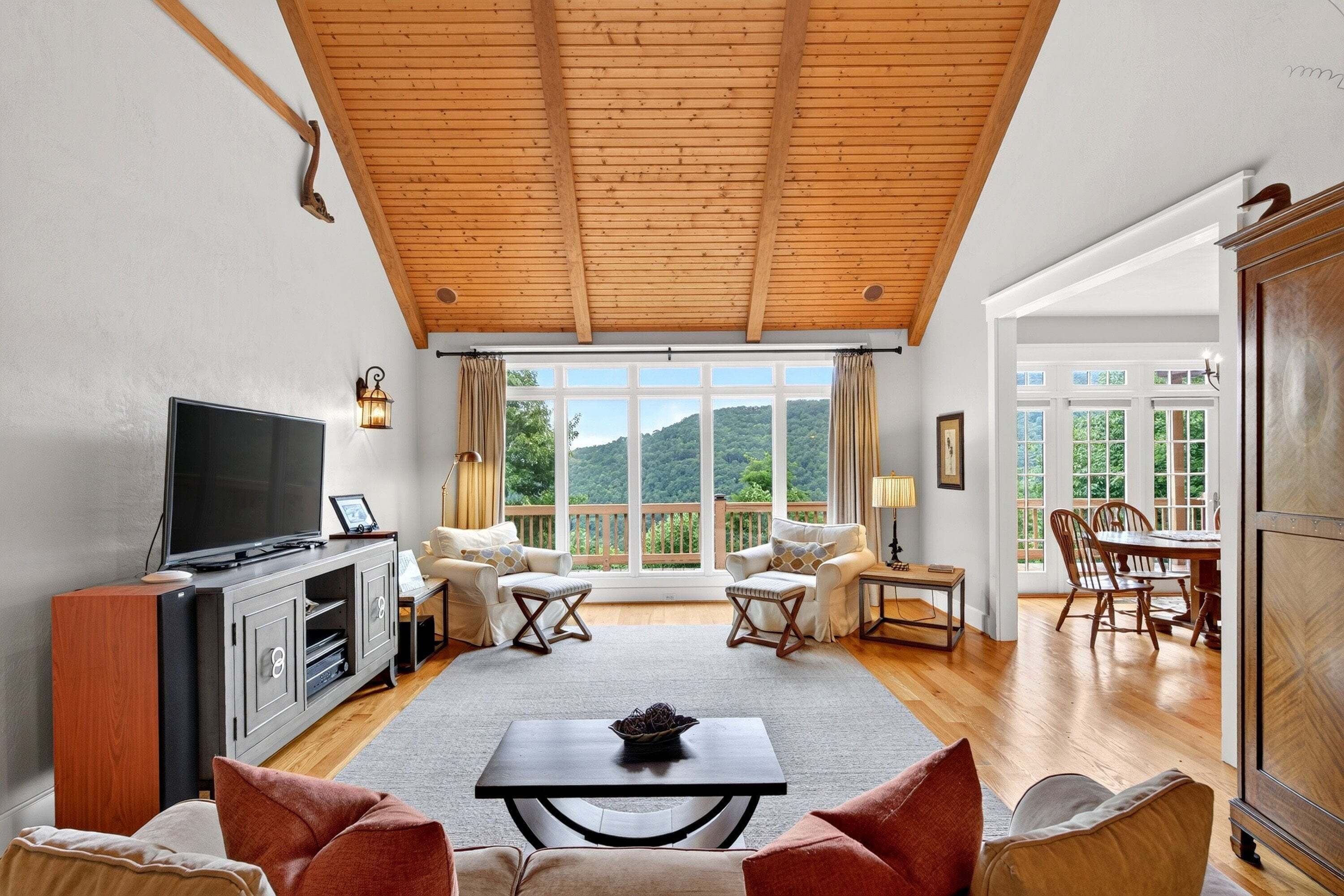 A living room with a TV, sofa, chairs, a dining table, and a large window with a view of greenery.