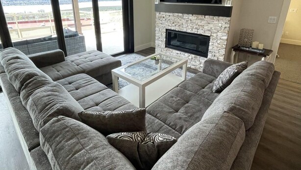 A modern living room with a large sectional sofa, a flat-screen TV mounted above a stone fireplace, and a ceiling fan. - Beautiful Private Residence at Spirit Ridge  (Osoyoos)