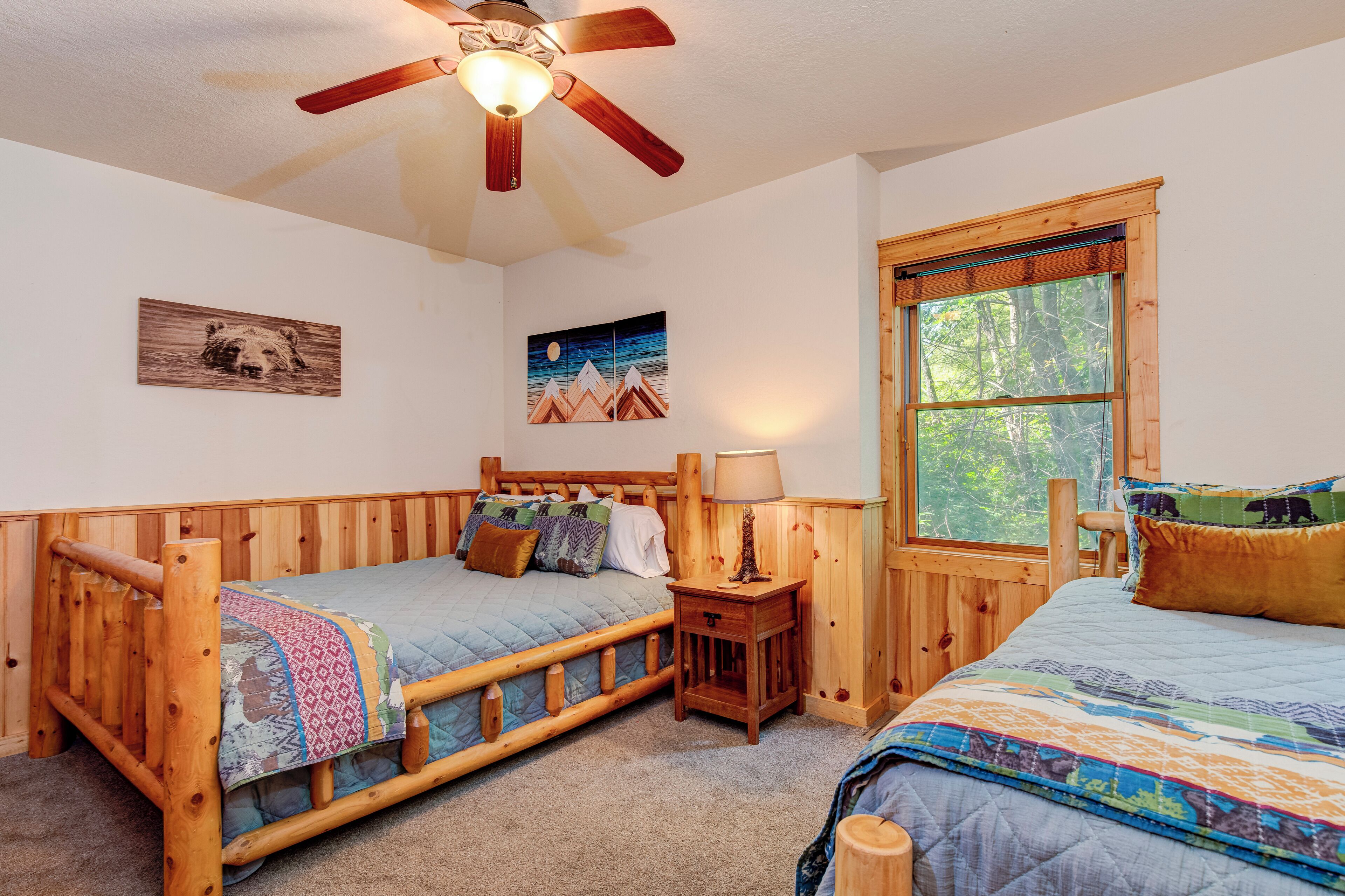 A cozy bedroom with two wooden bunk beds, a ceiling fan, a window with a view of trees, and wall art.