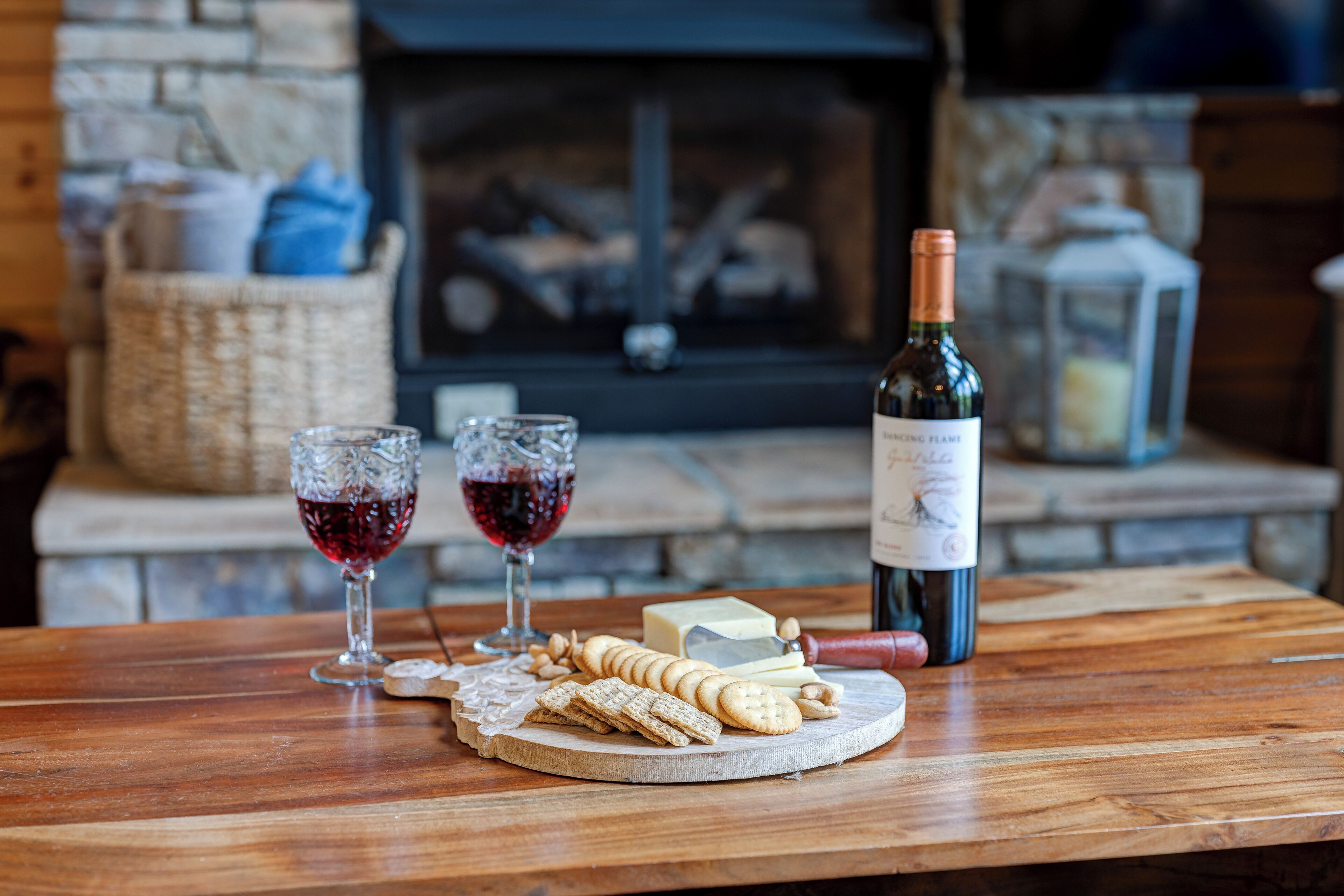A wooden table with a cheese board, crackers, and wine glasses. A bottle of wine is also visible.