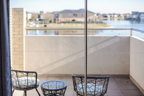 A balcony with two wicker chairs and a small round table, overlooking a body of water and buildings in the distance. - StayAU Magnificent Lakefront Villa  (Point Cook)