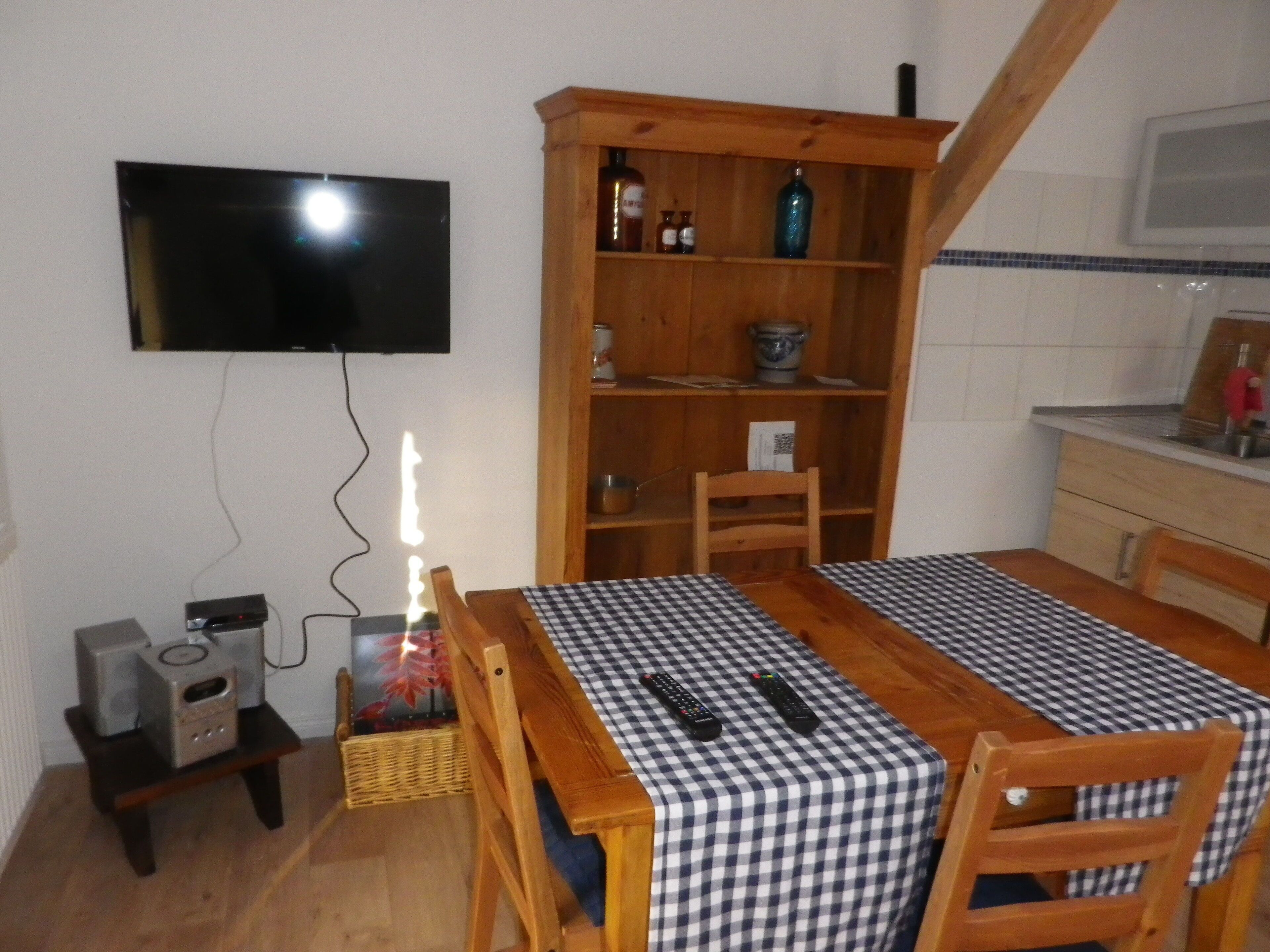 Un comedor con mantel a cuadros, sillas de madera, un televisor montado en la pared, una estantería de madera y una pequeña zona de cocina con microondas.