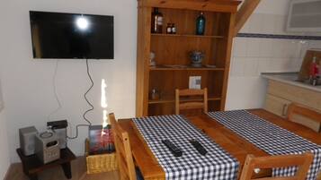 Un comedor con mantel a cuadros, sillas de madera, un televisor montado en la pared, una estantería de madera y una pequeña zona de cocina con microondas.