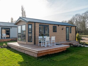 A modern, single-story house with a wooden exterior and a tiled roof. It features a deck with outdoor furniture and a large window. - Admirable Chalet Near the River (Kerkdriel)