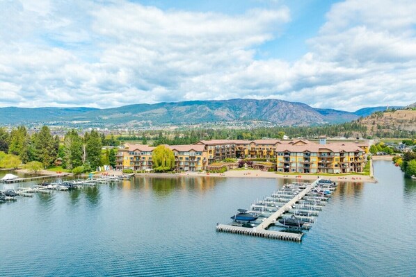 Hotel frente a un lago con un puerto deportivo, rodeado de montañas y vegetación.