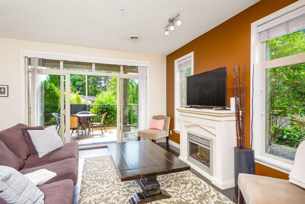 A living room with a sofa, coffee table, fireplace, TV, and a sliding glass door leading to a patio with outdoor furniture.