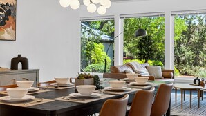 Un'elegante zona pranzo con tavolo in legno scuro apparecchiato per un pasto, circondato da poltrone in pelle. Ampie finestre offrono vista su un rigoglioso spazio verde.