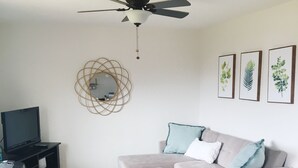 Un salon avec un canapé gris, un pouf blanc, un ventilateur de plafond, un téléviseur posé sur un meuble et des reproductions botaniques encadrées accrochées au mur.