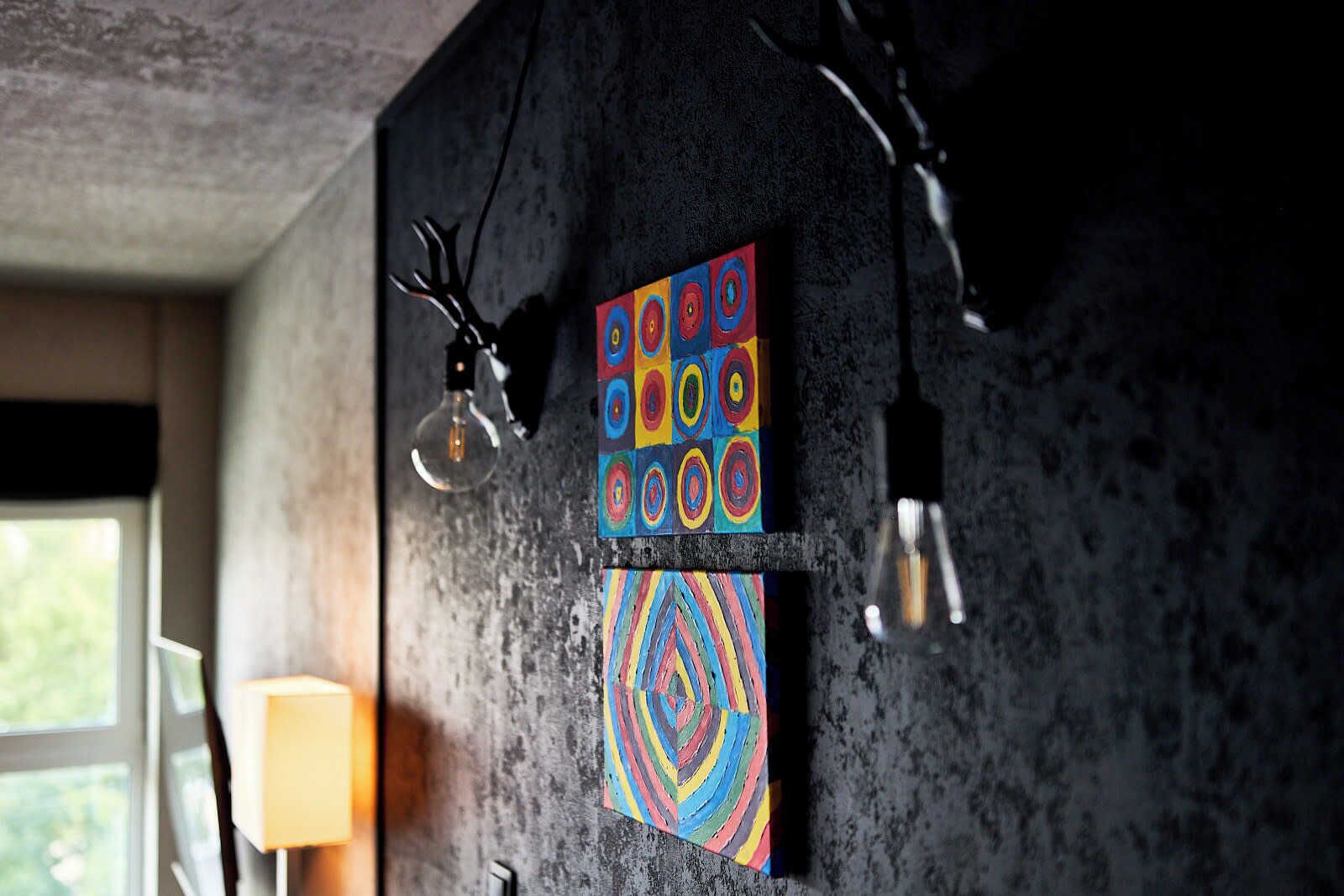 Ein Zimmer mit einer Betonwand, zwei farbenfrohen Bildern und einer Lampe.