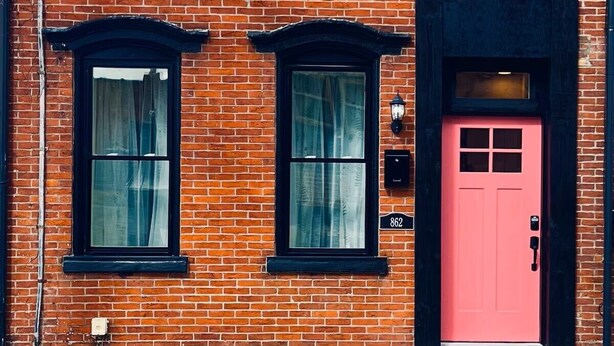 A red brick building with two black-framed windows and a pink door, featuring a small porch and a visible address number. - Cozy, walkable 1-bedroom apartment in Pittsburgh with WiFi, AC (Pittsburgh)
