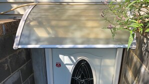 Eine kleine, weiße Struktur mit einer Einzel-Tür, einem Fenstergitter aus Metall, einem roten Aufkleber und einem Metallrost an der Basis.