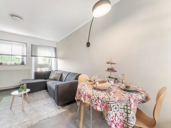 A modern living room with a grey sofa, a small round table, and a dining table with a colorful tablecloth. There is a window with blinds and a radiator on the wall. - Holiday Home in Voerendaal With Terrace (Voerendaal)
