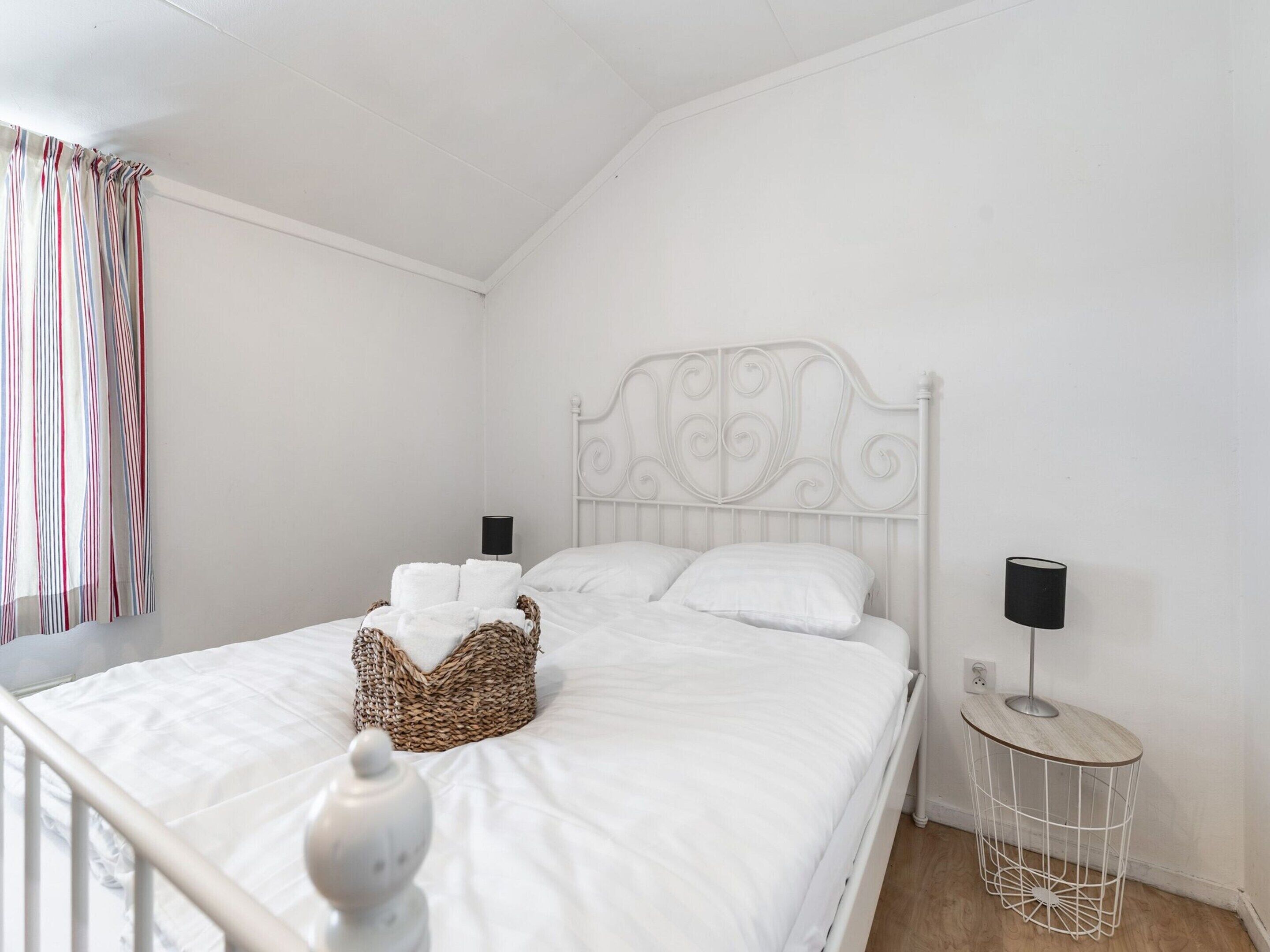 Een witte slaapkamer met een metalen hoofdbord, een nachtkastje met een lamp, en een rieten mand op het bed.