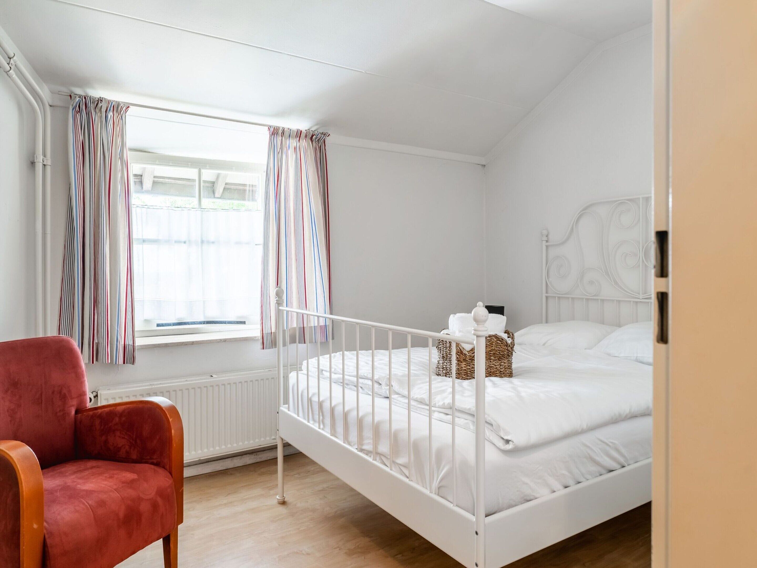 A bedroom with a white bed, a red chair, and a window with striped curtains.