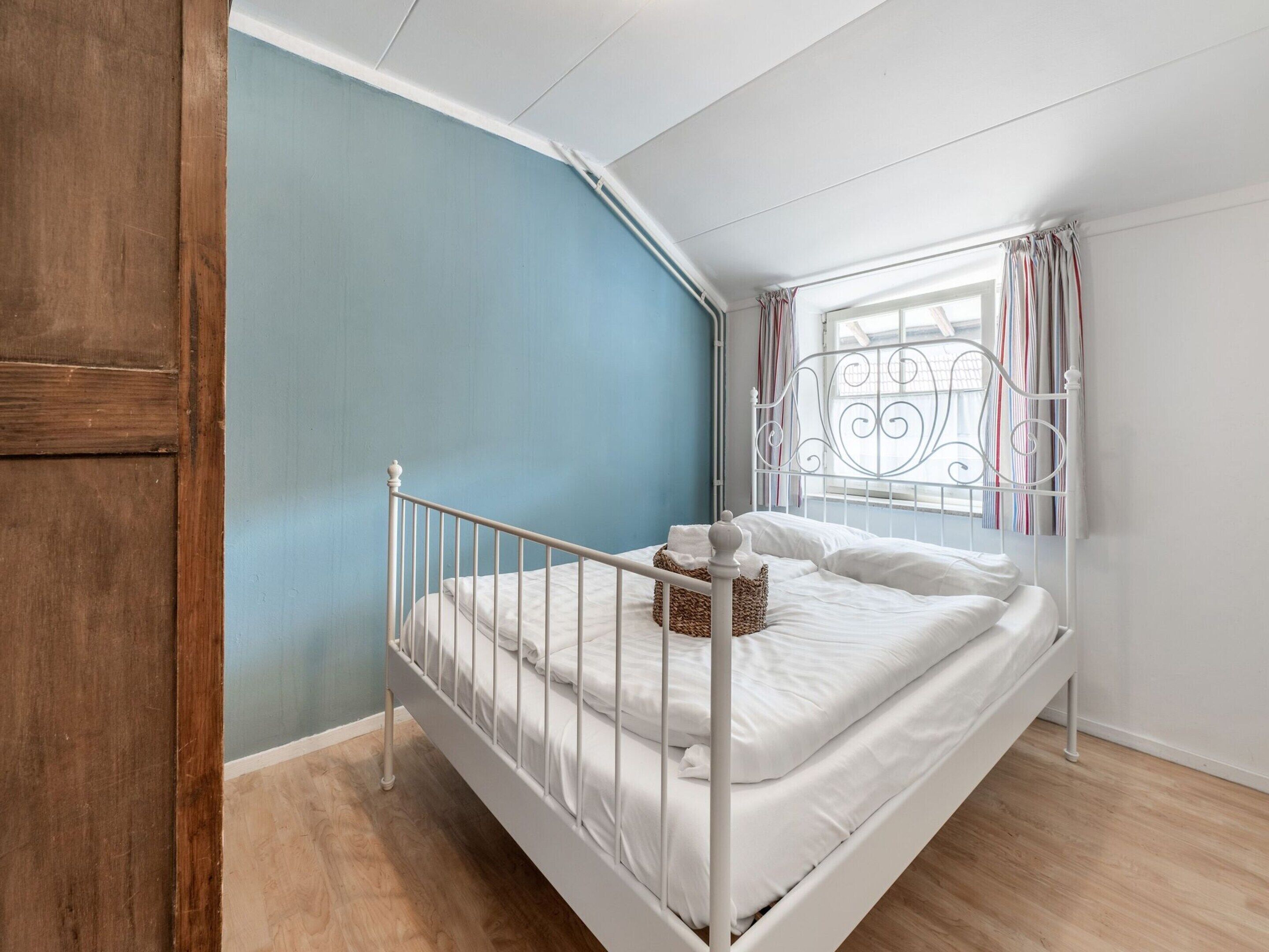 Une chambre avec un lit en métal blanc, un mur bleu, un plancher en bois et une fenêtre avec des rideaux.