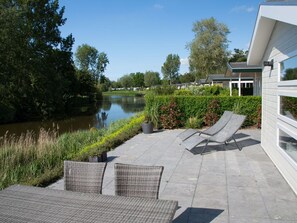 A patio with wicker furniture overlooking a calm river, surrounded by lush greenery and modern buildings. - Nice Tent With Bathroom, Near a Recreation Area (Noord-Scharwoude)