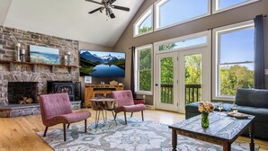 A spacious living room with a stone fireplace, comfortable seating, a television, and large windows offering a view of the outdoors. - Stunning Views, Riverfront Privacy, 18-Acre Estate - Near Hikes, Wine/Breweries! (Charles Town)