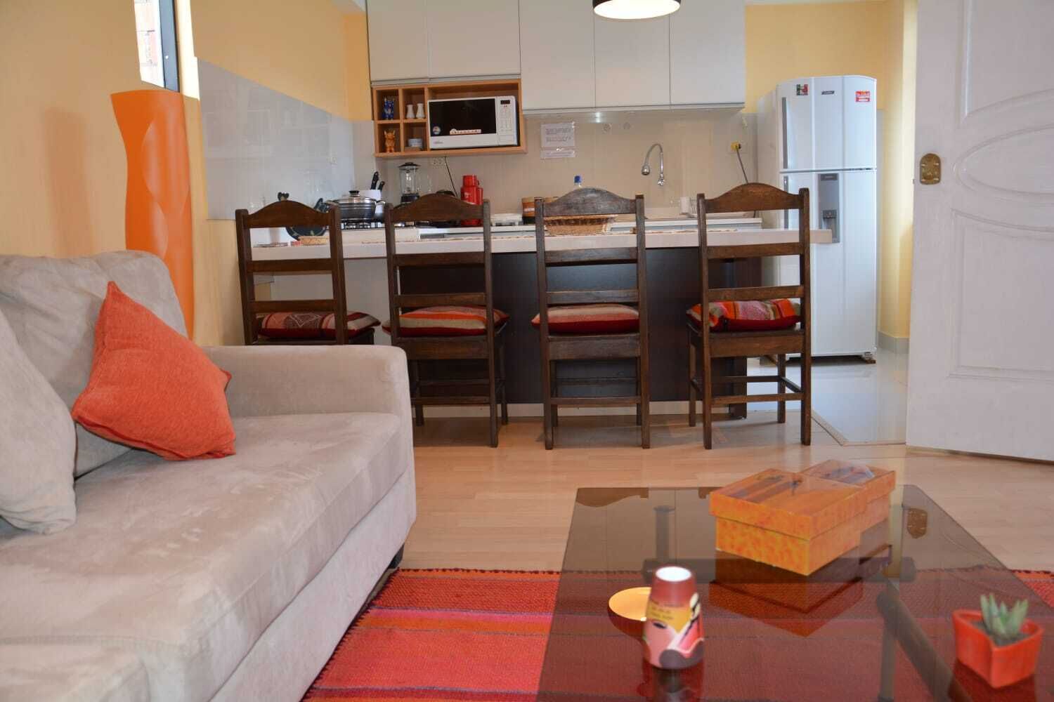A modern living room with a sofa, a coffee table, and a kitchen area with a microwave, refrigerator, and dining chairs.