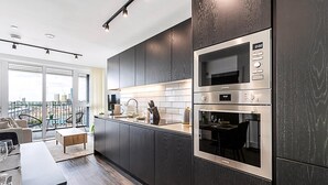 A modern kitchen with dark wood cabinets, stainless steel appliances, and a city view through large windows. - Sky-High Studio with Stunning Views (London)