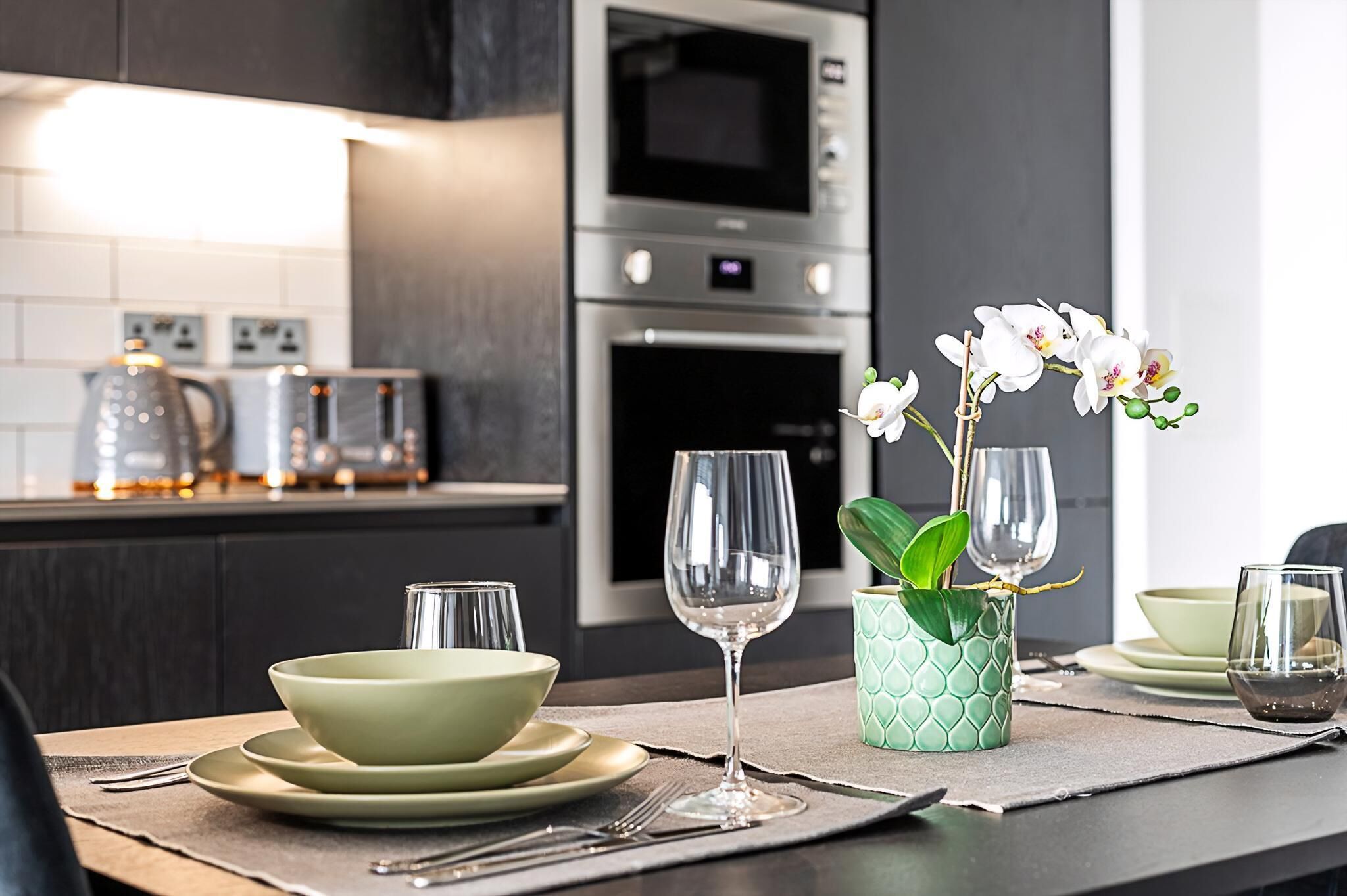 A modern kitchen with a dining table set for two, featuring a green bowl, wine glasses, and a vase with white flowers.