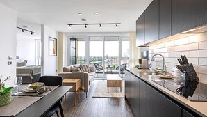 A modern kitchen with dark cabinetry, a dining area with a table and chairs, and a view of the city through large windows. - Sky-High Studio with Stunning Views (London)