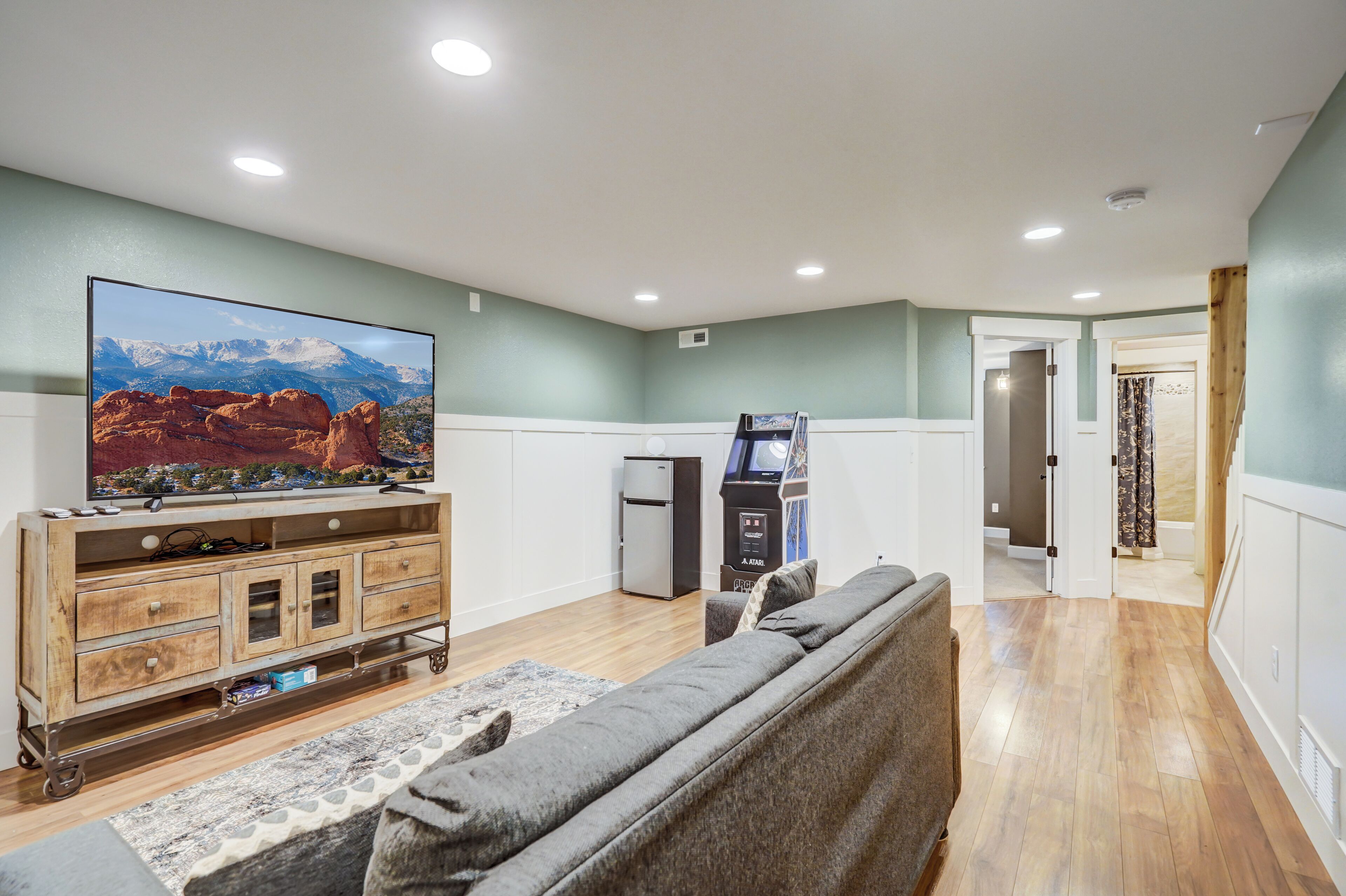 A living room with a large flat-screen TV, a wooden cabinet, a gray sofa, and a small refrigerator.