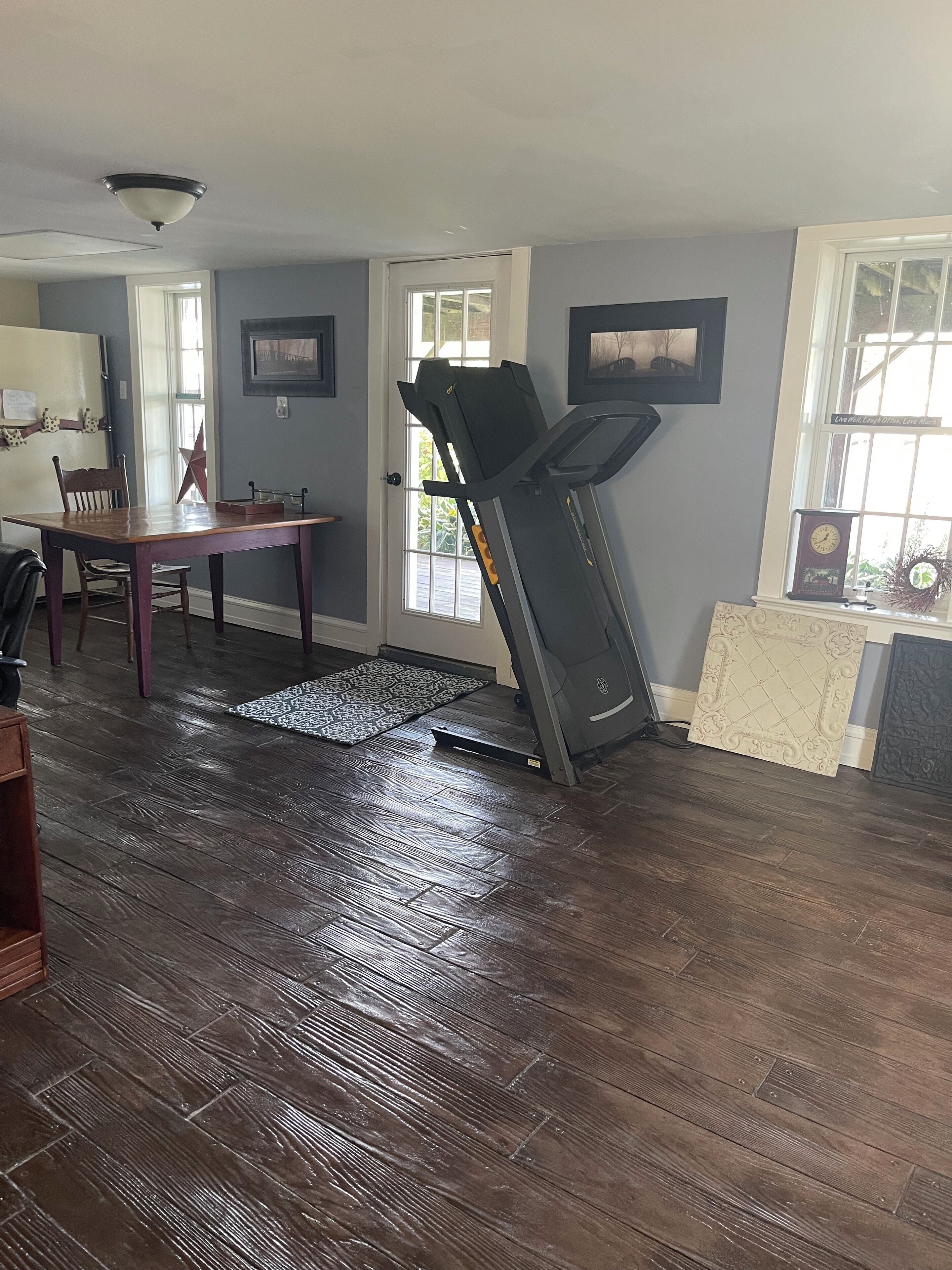 A room with a treadmill, a dining table, chairs, a rug, and a window with a wreath.