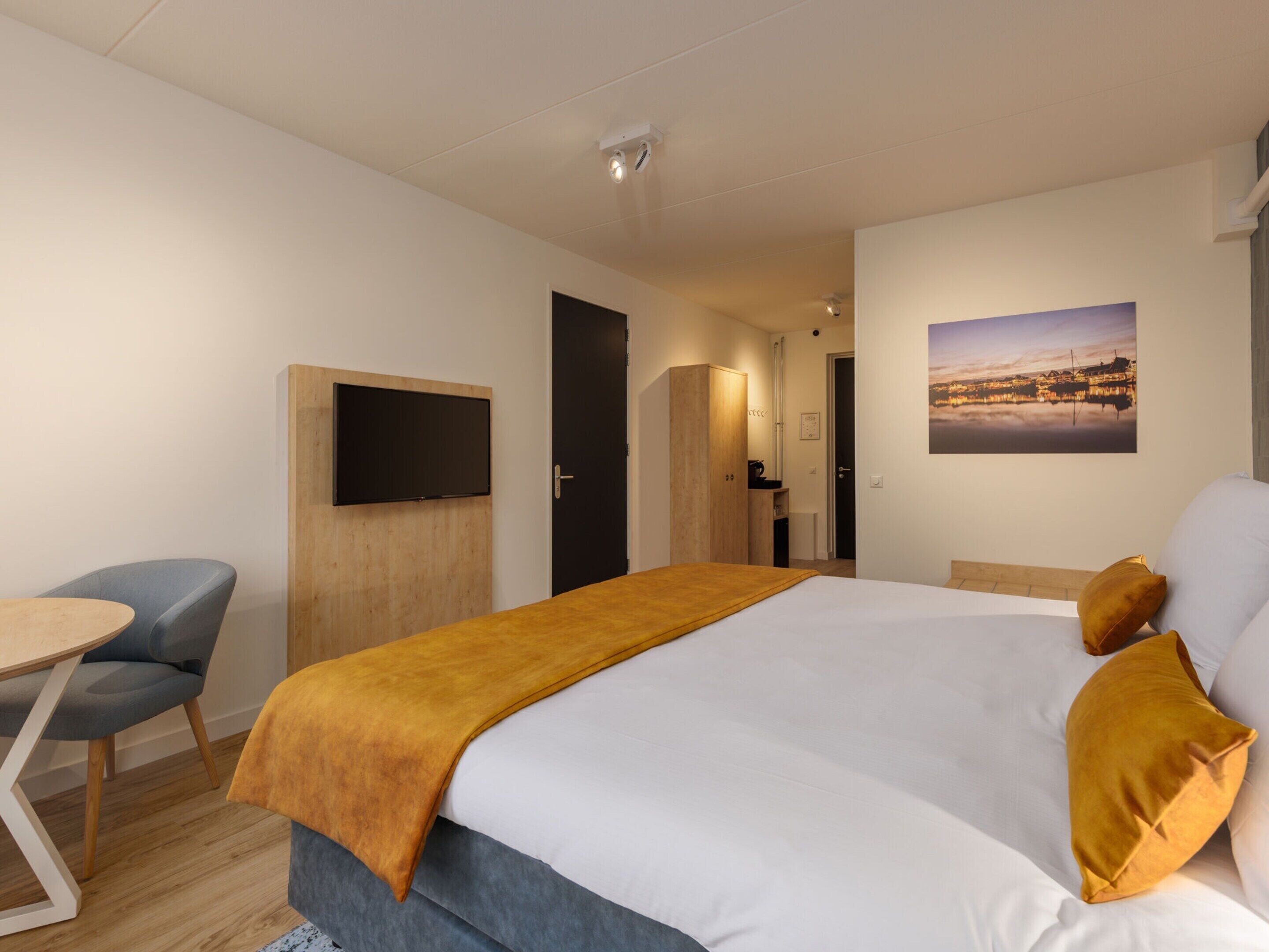 A hotel room with a bed, a desk with a chair, a TV mounted on the wall, and a painting on the wall.