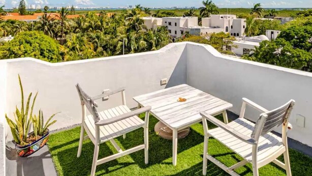 Eine Dachterrasse mit einem weißen Tisch und zwei Stühlen, umgeben von künstlichem Gras, mit Blick auf eine Küstenlandschaft mit Gebäuden und Palmen.