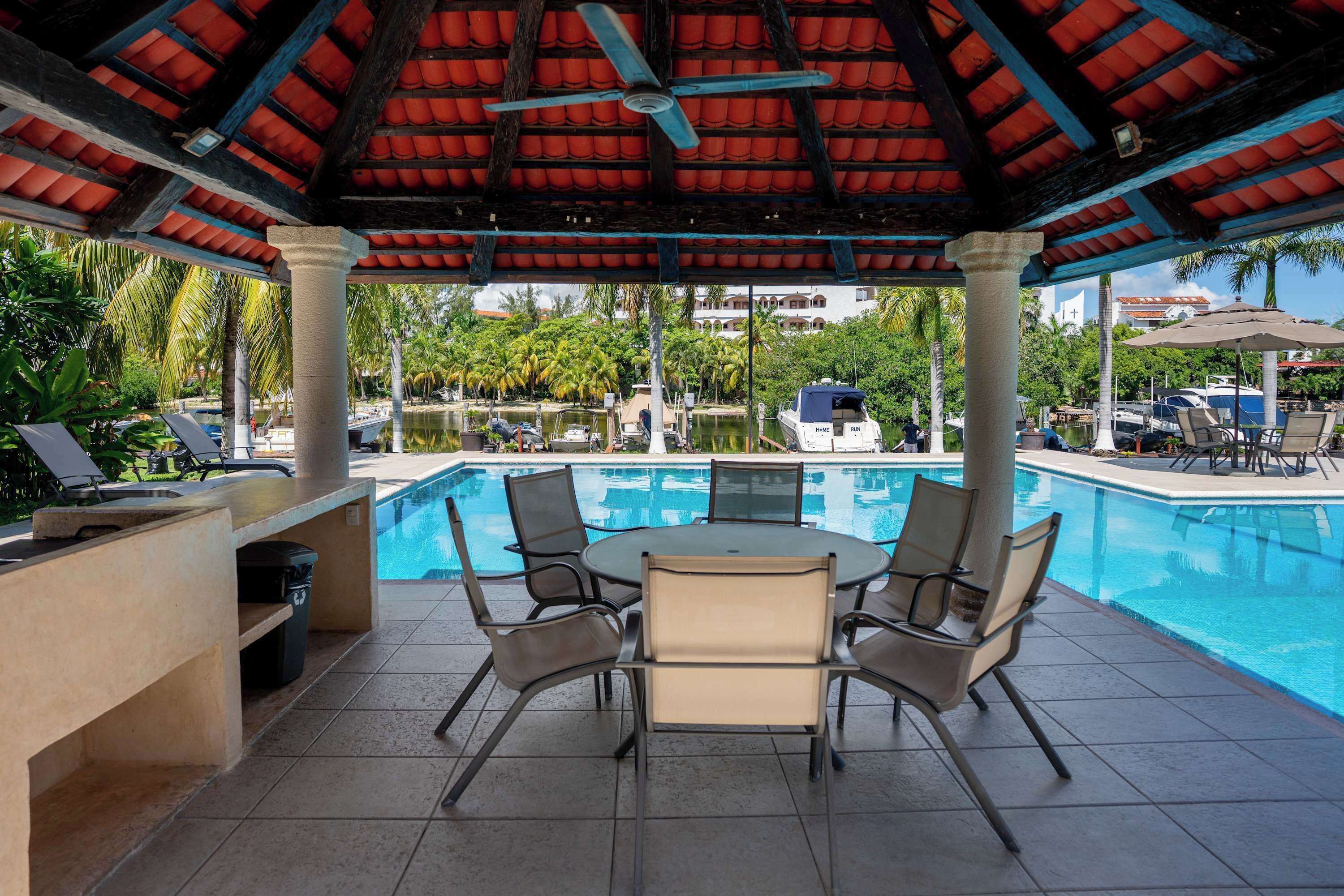 A covered outdoor pool area with a table and chairs, a ceiling fan, and a view of the pool and surrounding greenery.