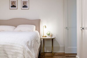 A neatly made bed with a white comforter and pillows, a bedside table with a lamp, and a door to the right.