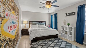 Ein Hotelzimmer mit einem Bett, einem Deckenventilator, einem Nachttisch, einer Lampe, einer Wanddekoration, einem Regal mit Dekorationen und einem Fenster mit Vorhängen.