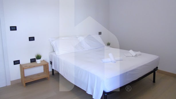 A neatly made bed with white linens, a small wooden nightstand with a plant, and a wall-mounted light fixture. - Homiday - Emma Residence (Giulianova)