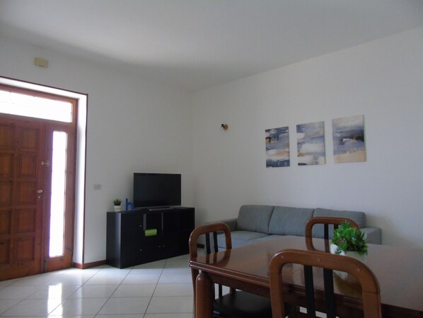 A living room with a sofa, a TV on a black stand, a dining table with chairs, and a door leading outside.