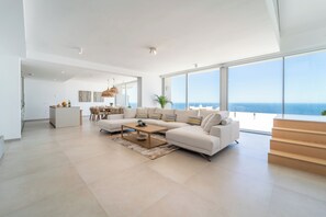 Ein geräumiges Wohnzimmer mit einem großen Fenster und Meerblick, eine moderne Küche im Hintergrund sowie eine Treppe, die zu einer oberen Etage führt.