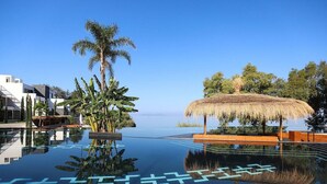Cielo azul despejado, una piscina infinita que refleja el paisaje, una estructura con techo de paja, palmeras y edificios modernos.