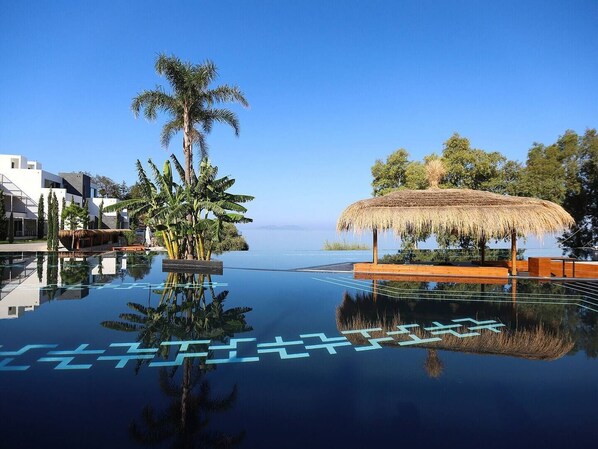 Cielo azul despejado, una piscina infinita que refleja el paisaje, una estructura con techo de paja, palmeras y edificios modernos.