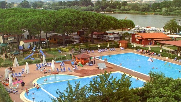 Piscina con sillas de descanso y sombrillas, rodeada de árboles y vista a un lago y colinas.