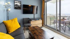 A modern living room with a sofa, a coffee table, a TV, and a view of the city through large windows. - Port Arcachon by Interhome (Arcachon)