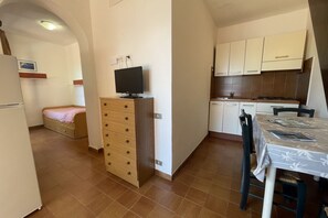 A compact kitchen with a dining area, a television on a wooden cabinet, and a bed with a pink blanket.