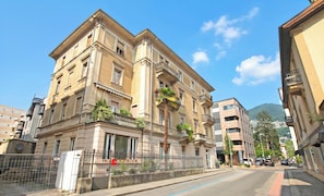 A multi-story building with balconies, a gated entrance, and a clear view of the street and mountains in the background.