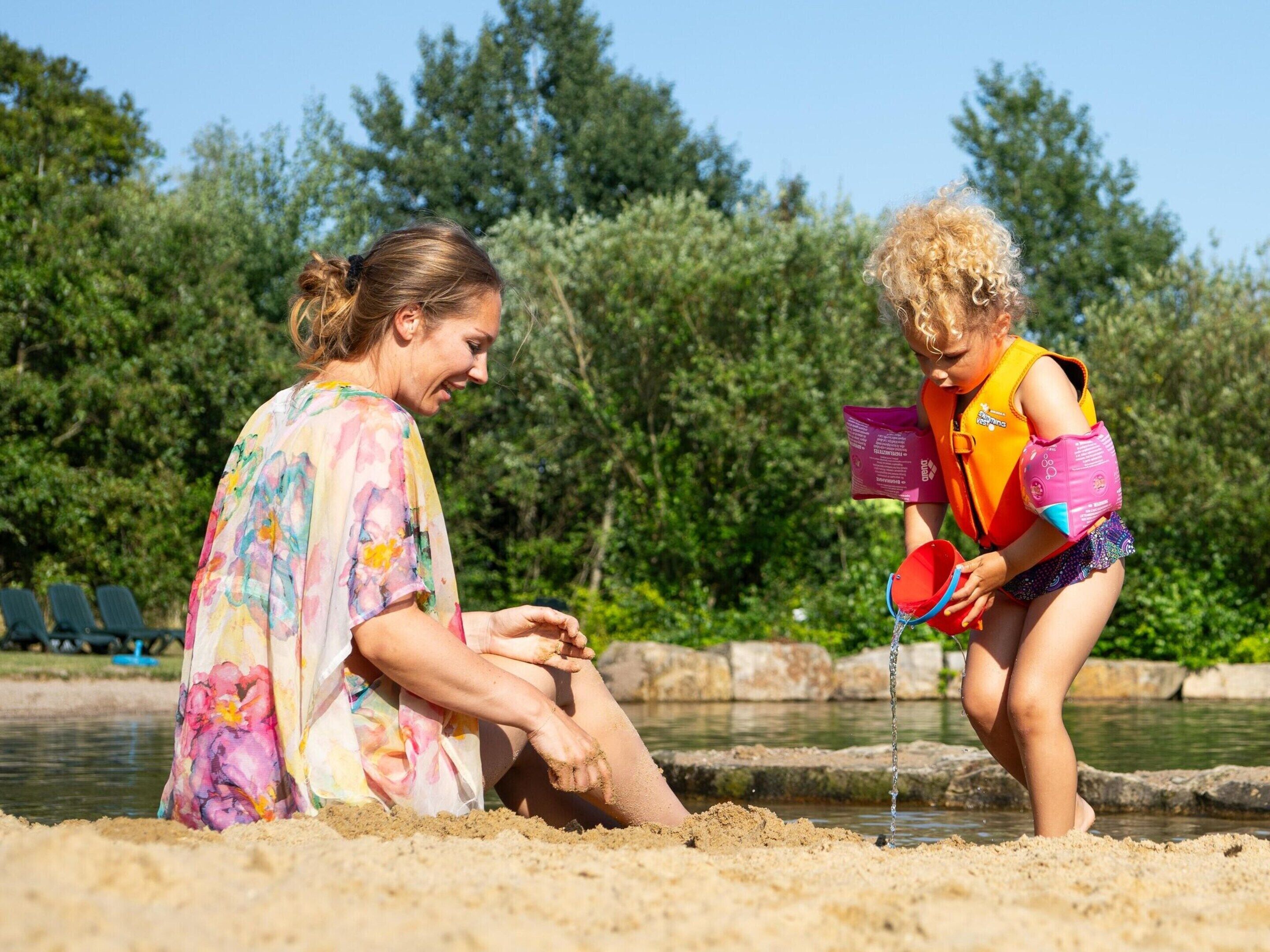 Two people playing in a sandy area near a water body, with greenery in the background.
