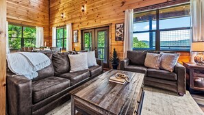 Interior acogedor de una cabaña con paredes de madera, dos sofás de cuero, una mesa de centro y grandes ventanales con vistas al bosque.