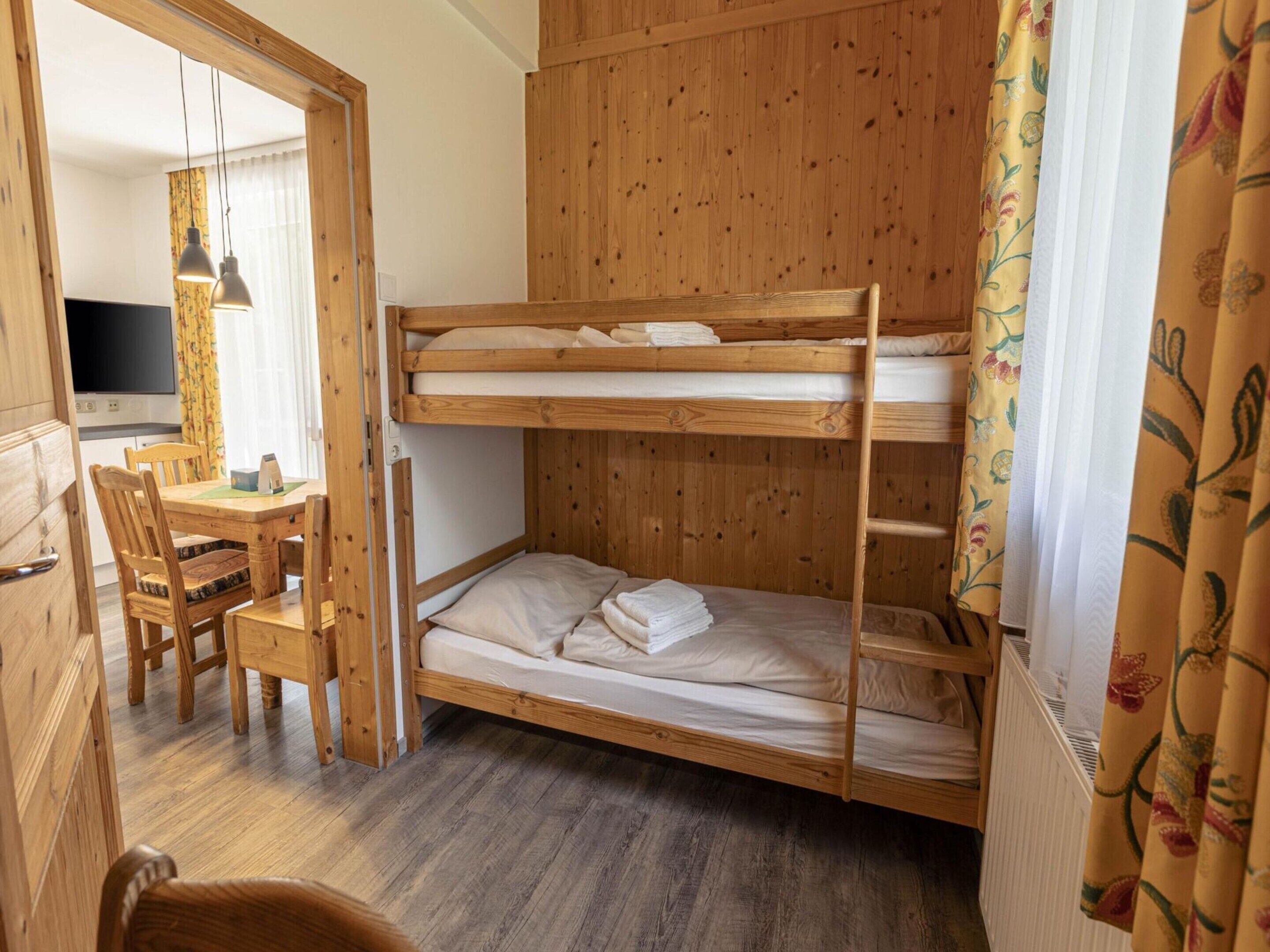 A bunk bed room with a wooden desk and chair, a television, and a window with curtains.