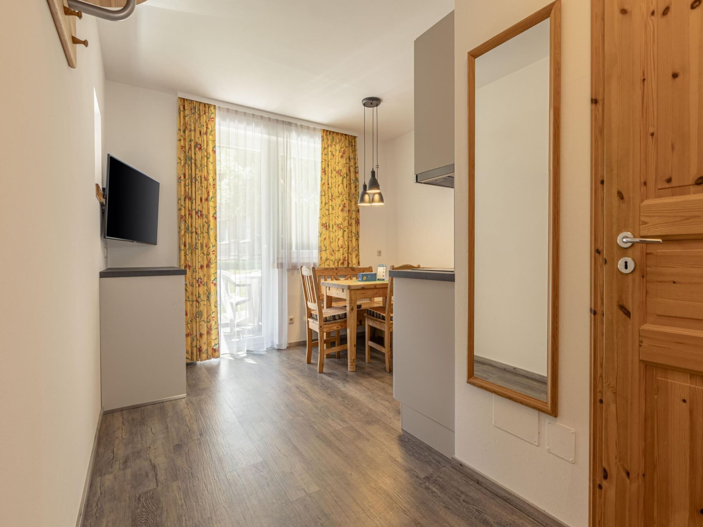 A compact room with a wooden door, a TV mounted on the wall, a dining table with chairs, and a window with curtains.