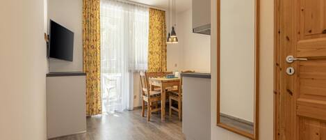 A compact room with a wooden door, a TV mounted on the wall, a dining table with chairs, and a window with curtains.