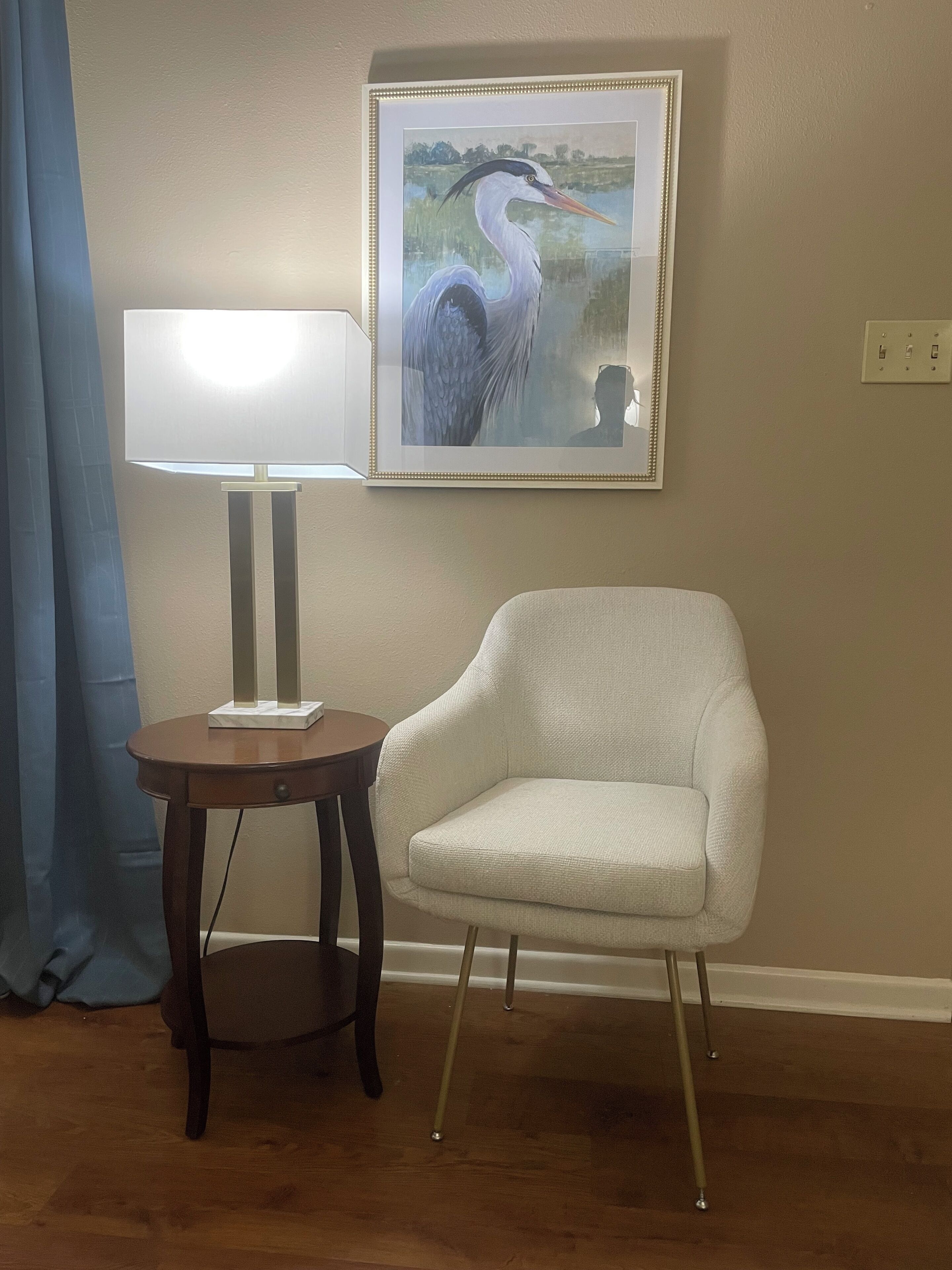 A room with a white armchair, a table with a lamp, and a framed picture on the wall.