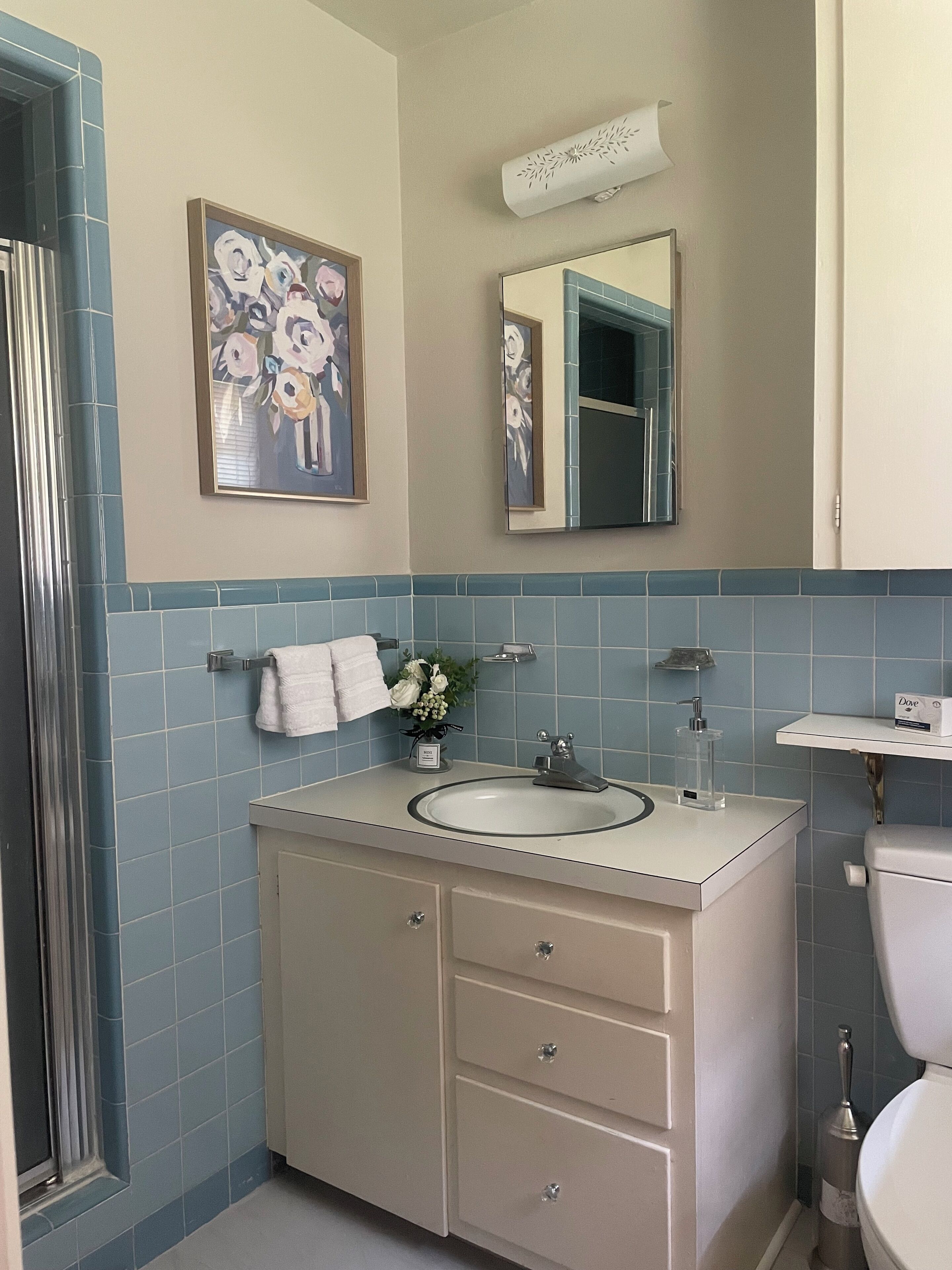 A bathroom with a sink, mirror, and toilet. There is a towel rack with a towel and a small vase with flowers. The walls are tiled in blue and white.