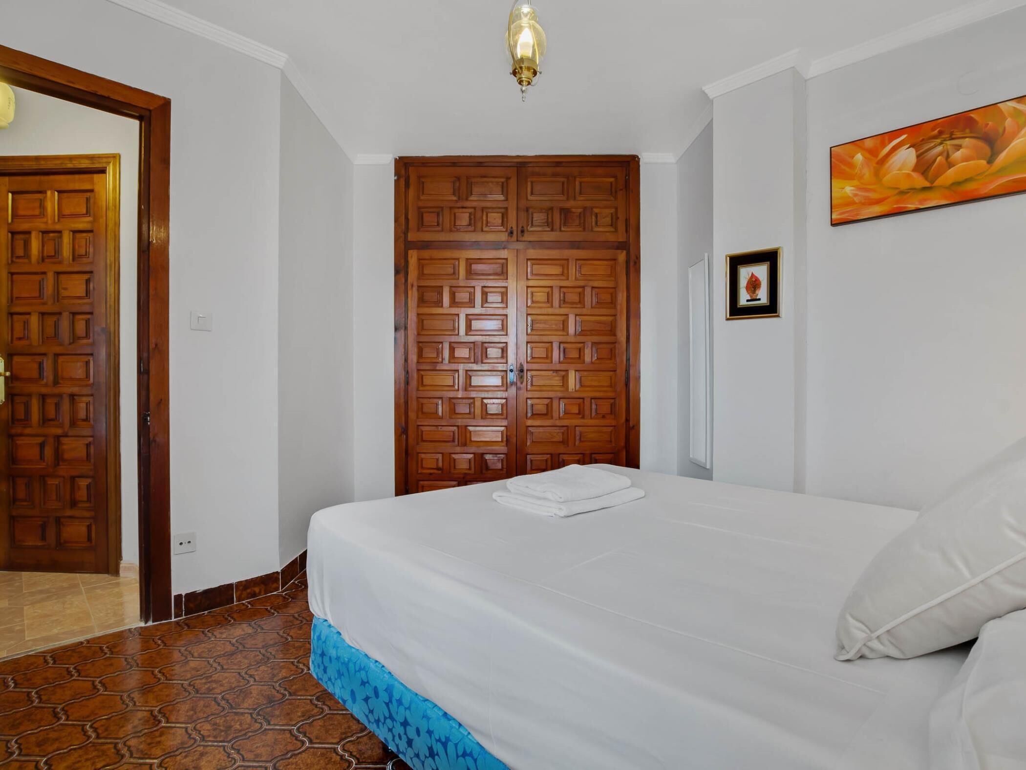 A bedroom with a bed, a wooden wardrobe, a painting on the wall, and a tiled floor.