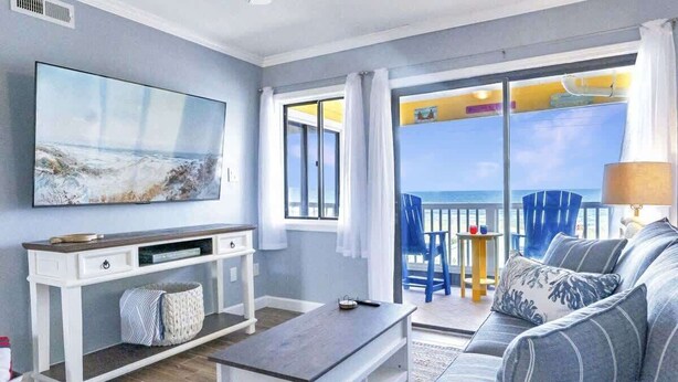 A living room with a large TV, a sofa, a coffee table, and a balcony with a view of the sea.